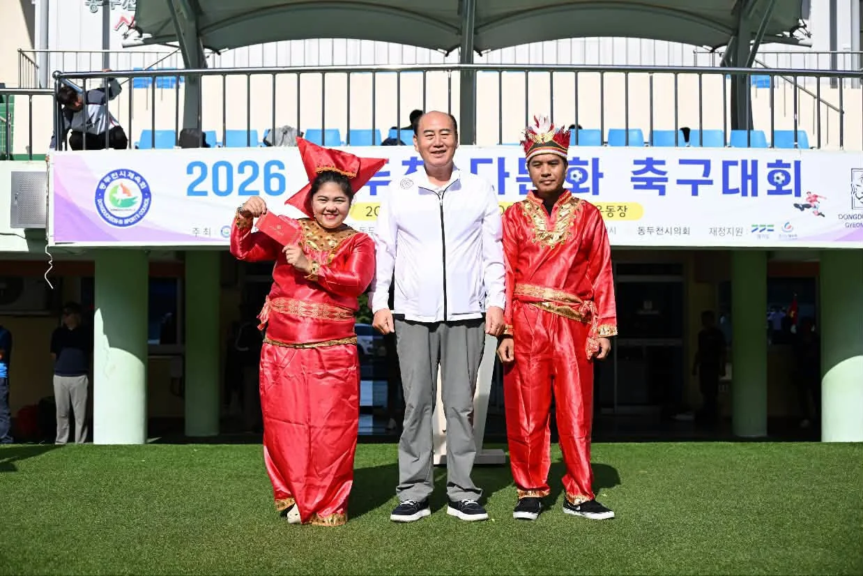 The tournament banner at 동두천종합운동장, with two of the Indonesian side in traditional dress. The banner names 동두천시체육회 as host, 동두천시 and 동두천시의회 alongside, and 경기도 as a financial supporter. Photograph by 박형덕, Mayor of Dongducheon.